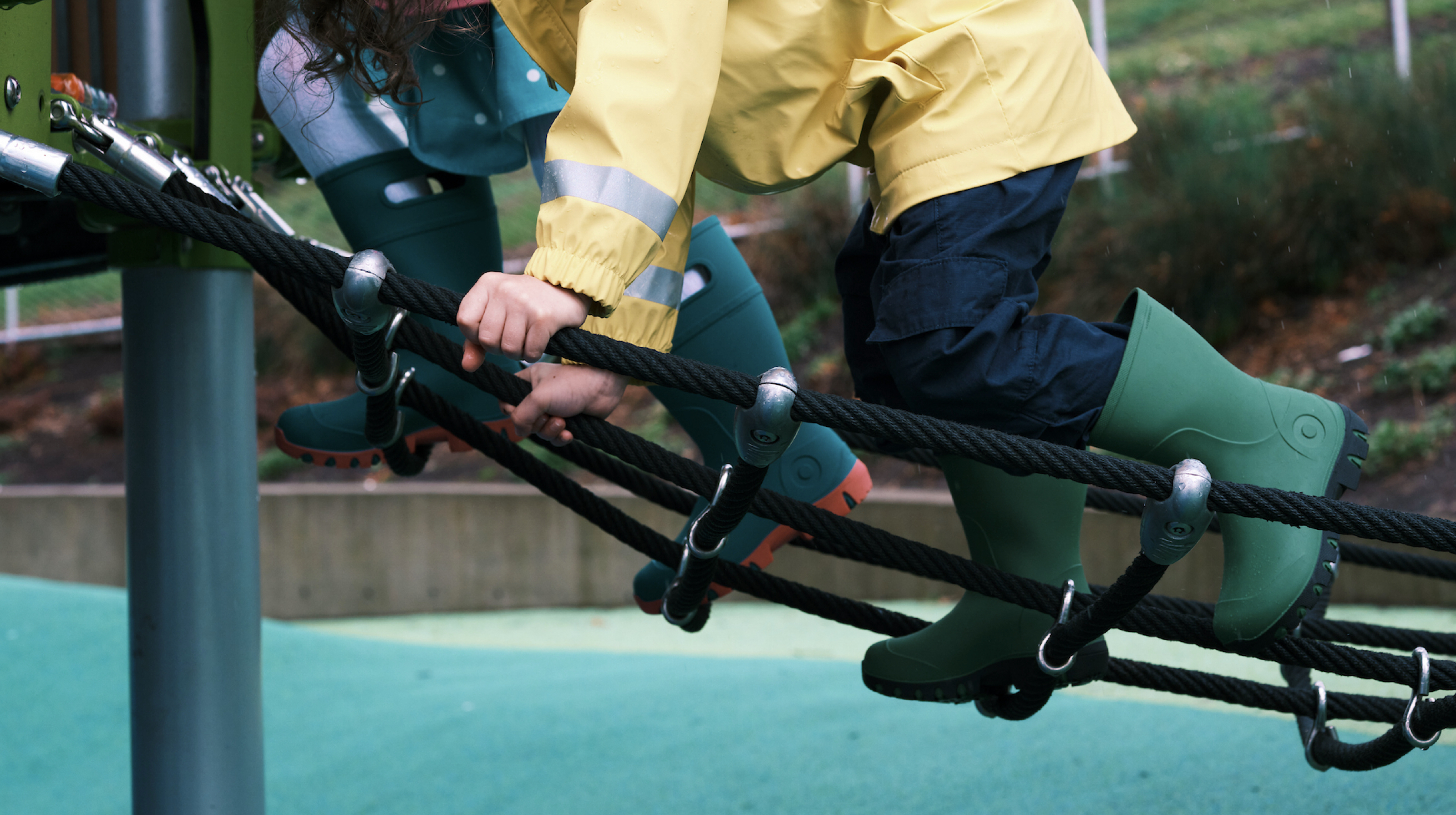 Two kids climbing up a rope ladder at a playground.  Wearing our Kids' Essential rain boots, shop now