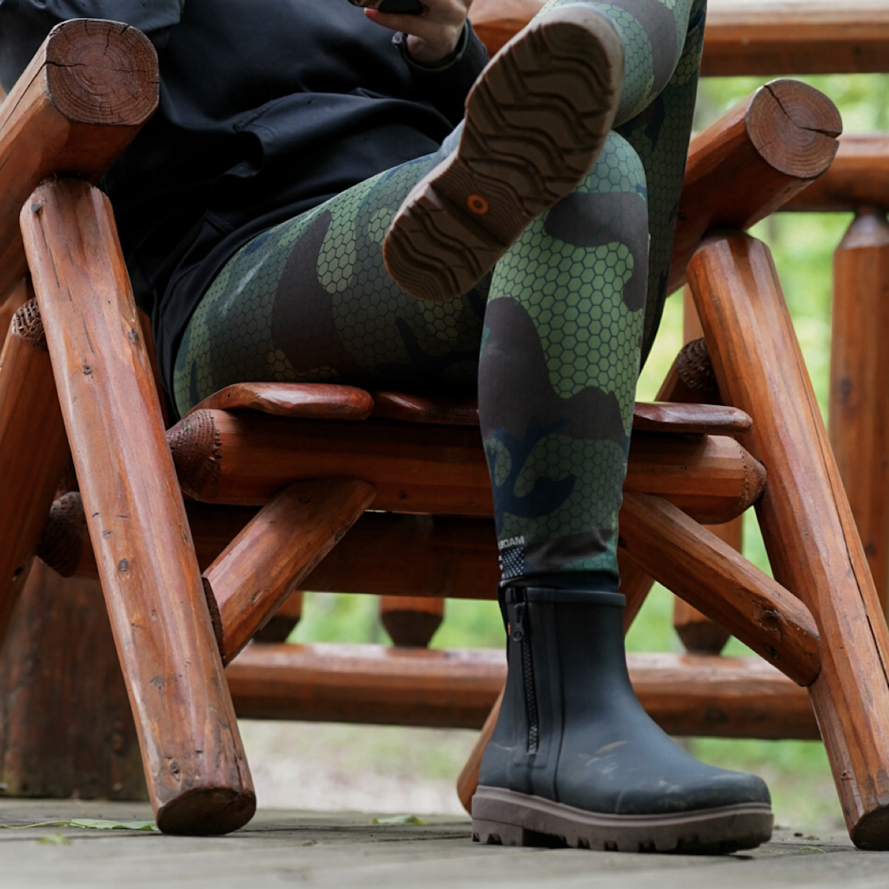A woman sitting down on a wooden lounge chair wearing the Holly Rain zip boots in black.