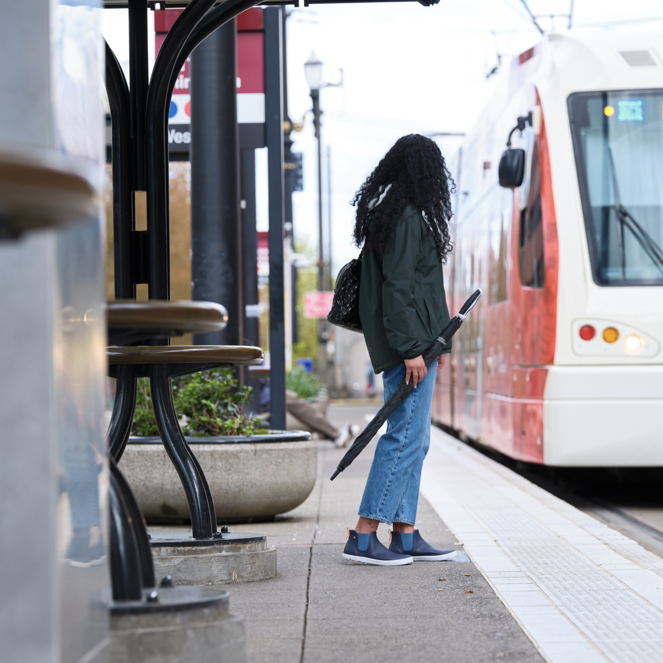 A woman waiting for the light rail wearing the Kicker Rain chelsea in blue holding an umbrella. 