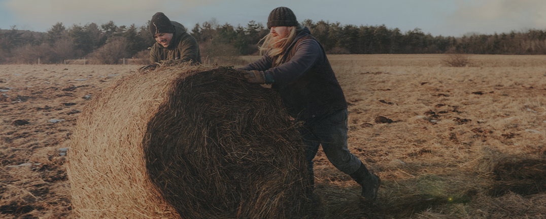 Shop our essentials for cold weather featuring 2 men rolling a haybale. 