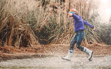 Image shows a sideways view of a kid                splashing around in a big puddle while wearing BOGS rainboots.