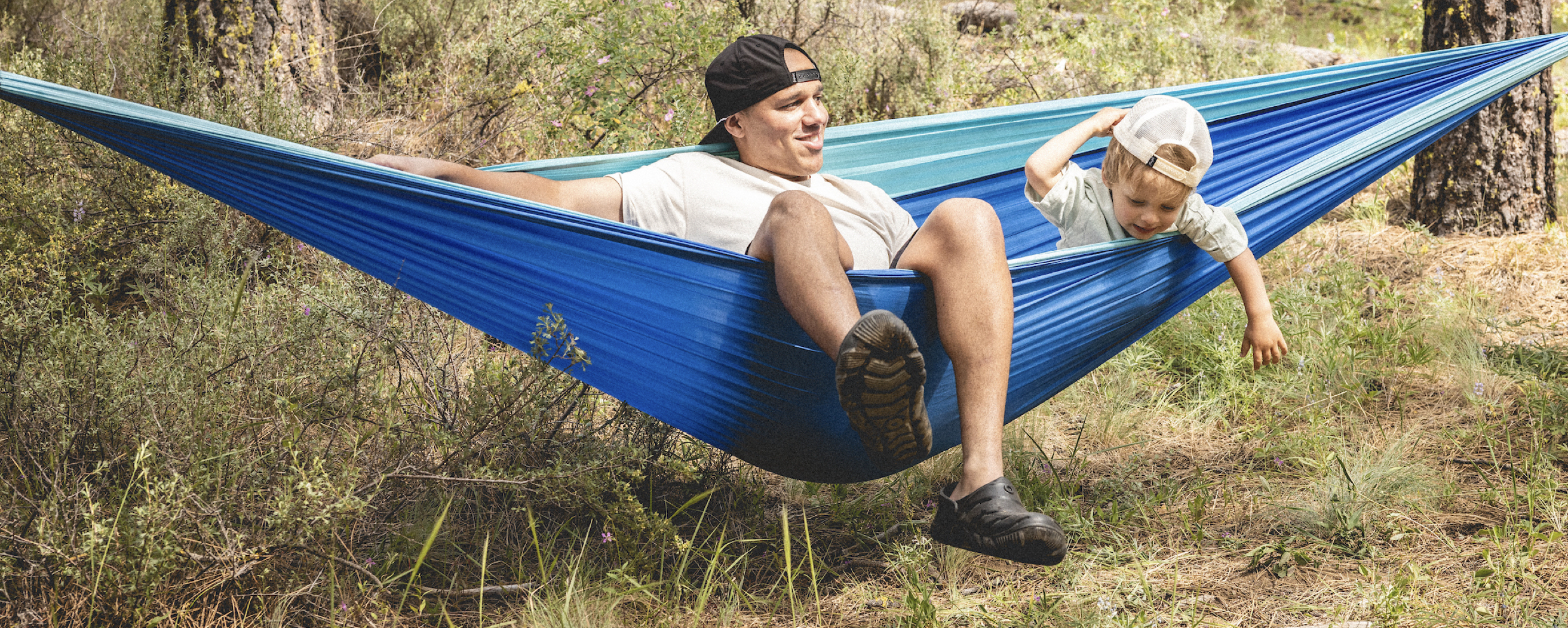 A man with his kid on a hammock.  Shop our Adult & Kids’ Boga casual slip on crocs. 