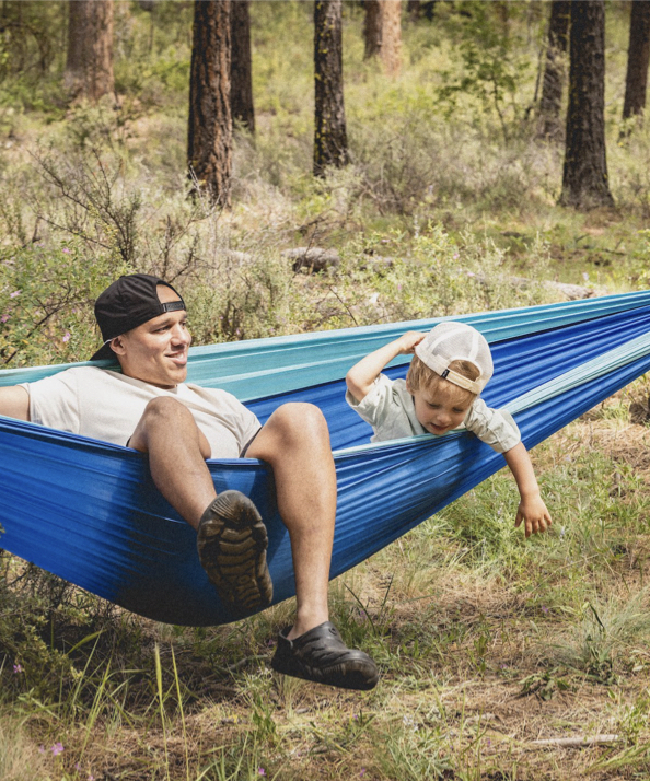 A man with his kid on a hammock.  Shop our Adult & Kids’ Boga casual slip on crocs. 