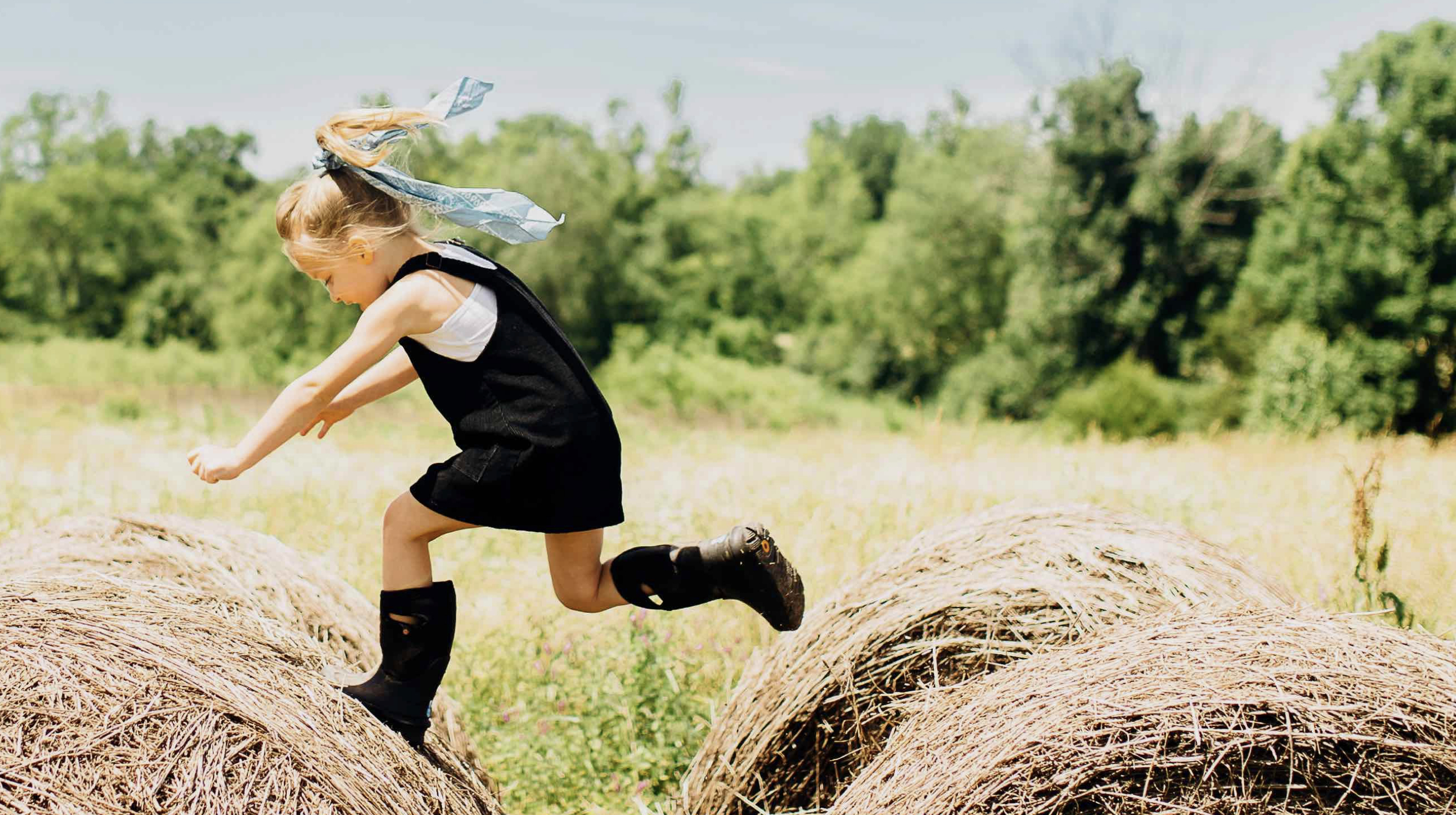 A girl jumping from one big hay barrel to another.  Shop our Kids' All-Season boots.