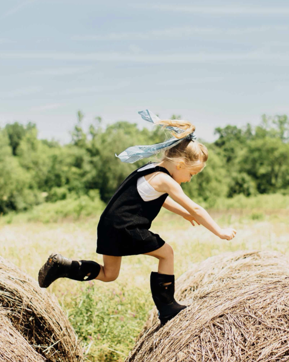 A girl jumping from one big hay barrel to another.  Shop our Kids' All-Season boots.
