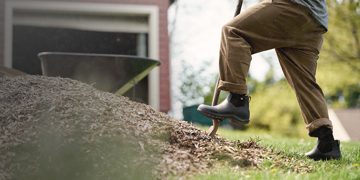 Image features a man in a black ankle boot shoveling mulch.