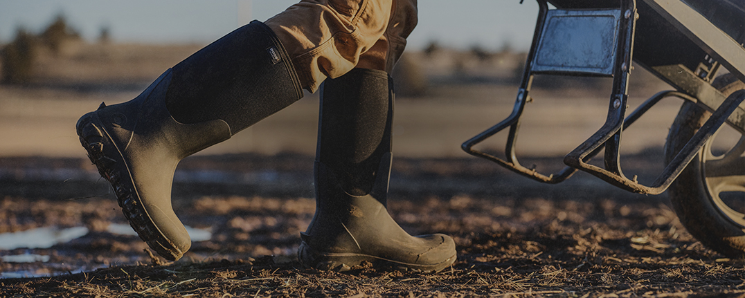 Image features a man walking in a muddy field wearing tall black boots. 