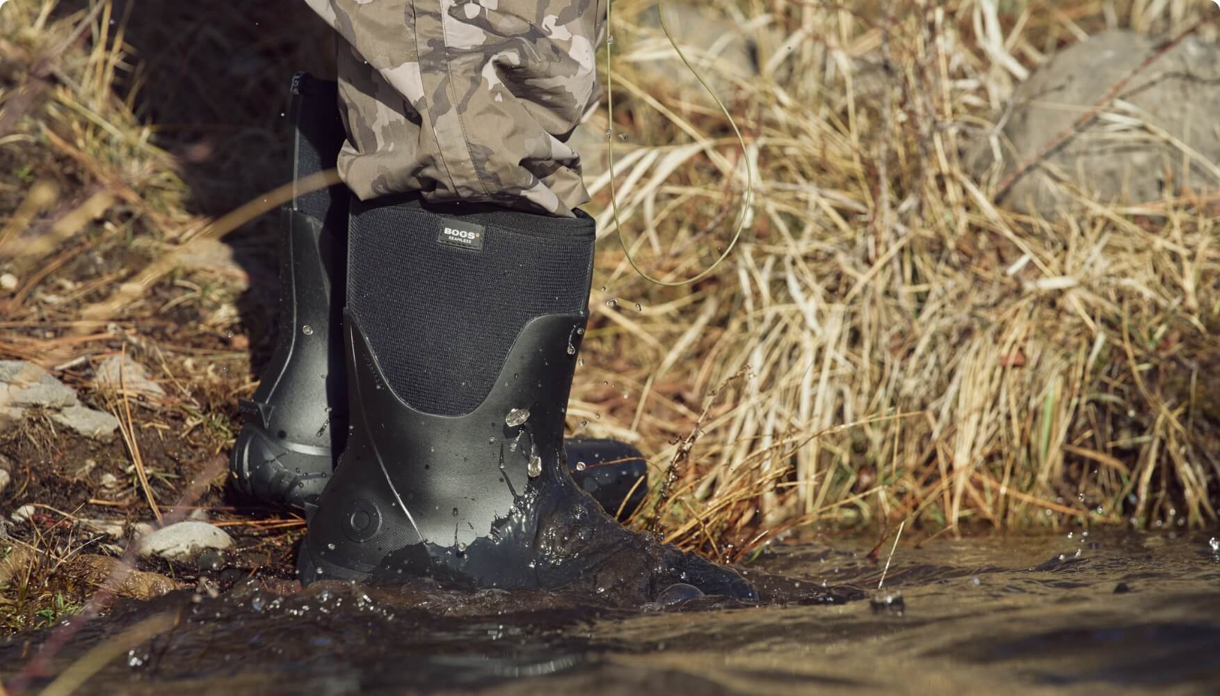 Image features BOGS Men's Insulated Waterproof Work Boots in Black being                   submerged in water next to dry grass.