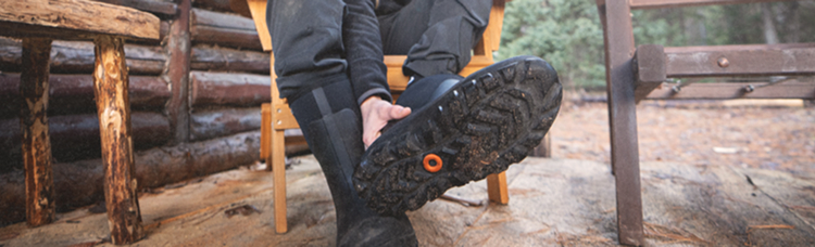 Image features a model removing their BOGS boot while sitting on a                chair outside of a cabin, highlighting the outsoles with aggressive tread and self-cleaning lugs.