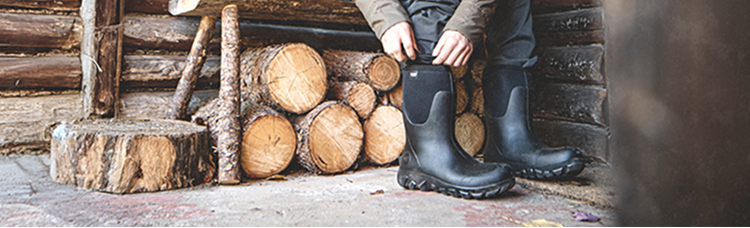 Image shows a distant shot of a model putting on BOGS                Seamless boots while sitting in front of cabin with a pile of logs.