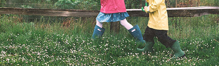 Image shows two kids in rain coats walking through a field                while wearing BOGS rain boots.
