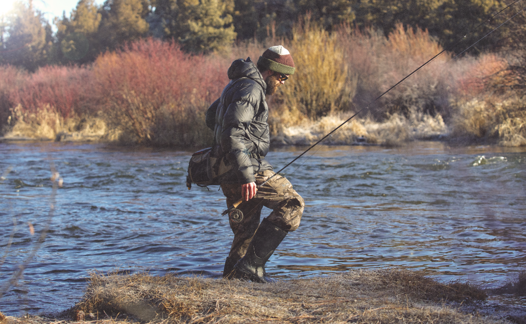 Image features a man fishing in a river on a sunny day. 